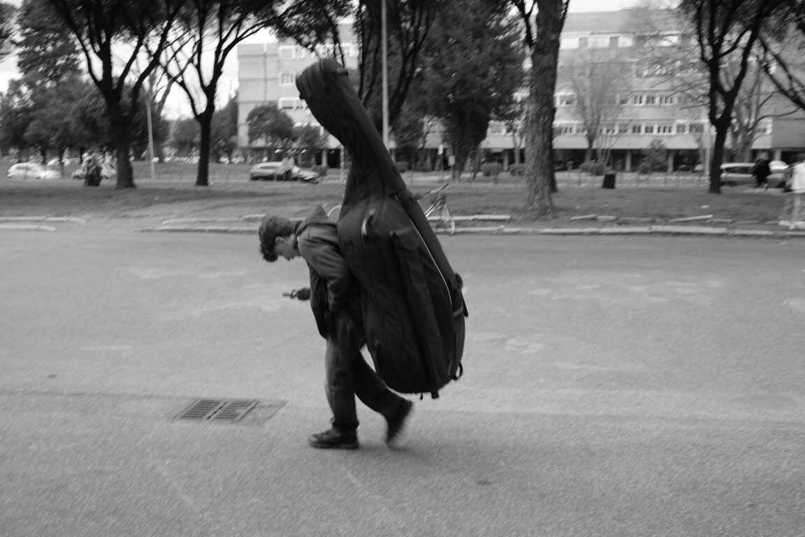 Man walking on a city street with a large contrabass case strapped to his back.