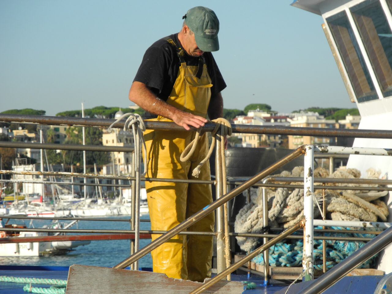 Harbor worker in yellow waterproof overalls on a boat deck handling ropes.