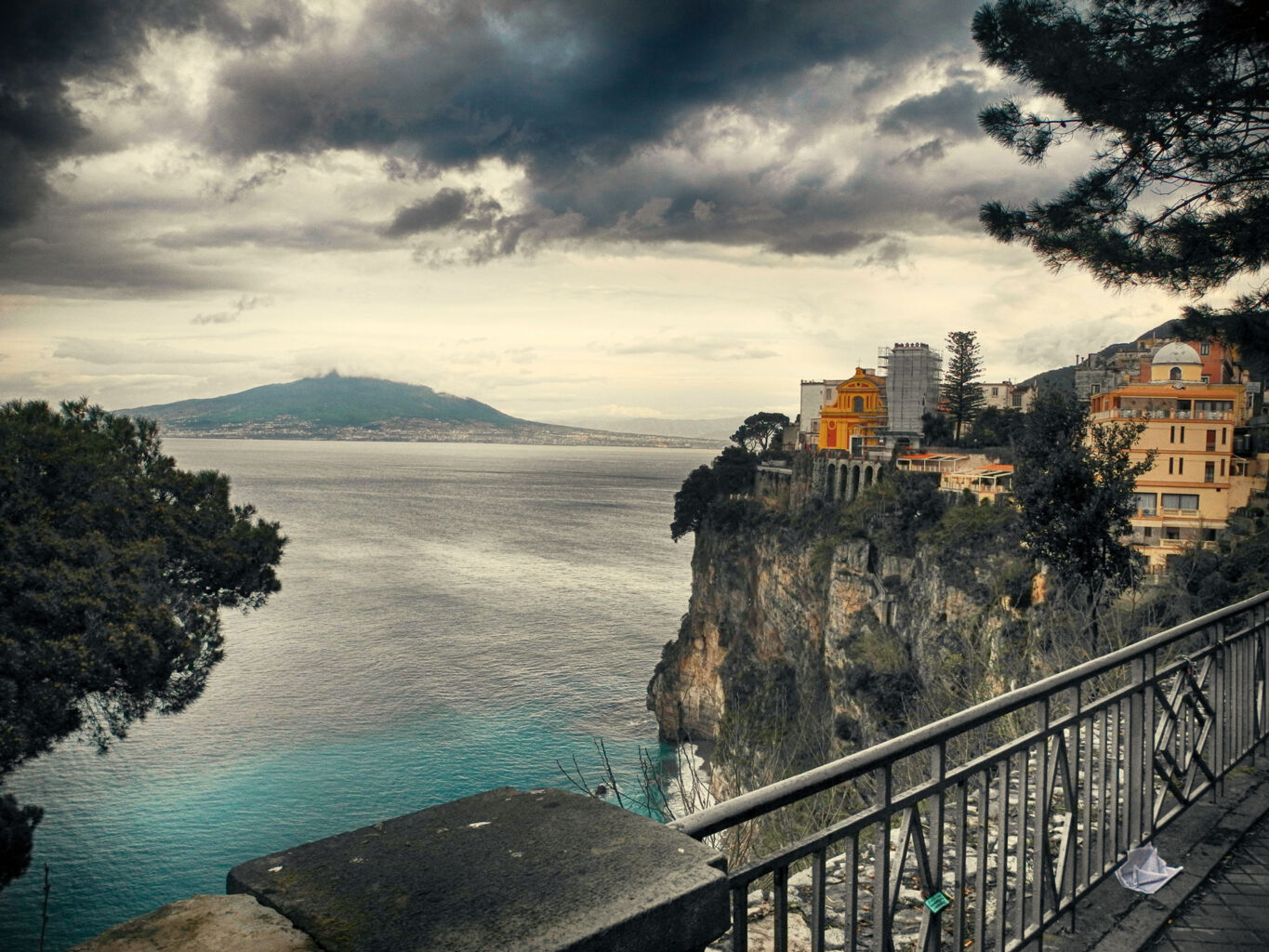 Veduta panoramica di scogliera con case colorate sul mare turchese, cielo nuvoloso e balaustra in primo piano