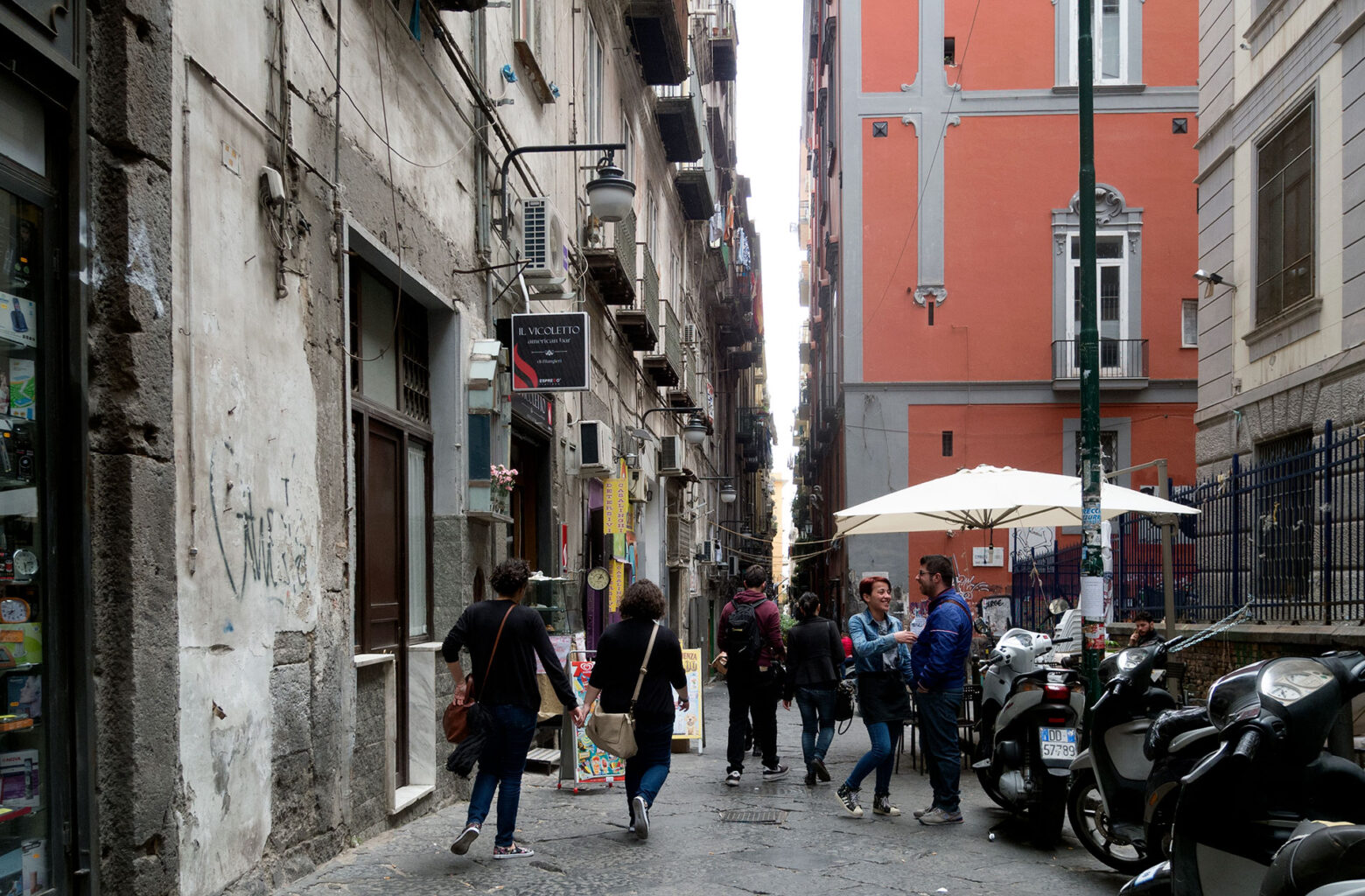 Vicolo nel centro storico con pedoni, scooter parcheggiati e tavoli all'aperto sotto ombrellone