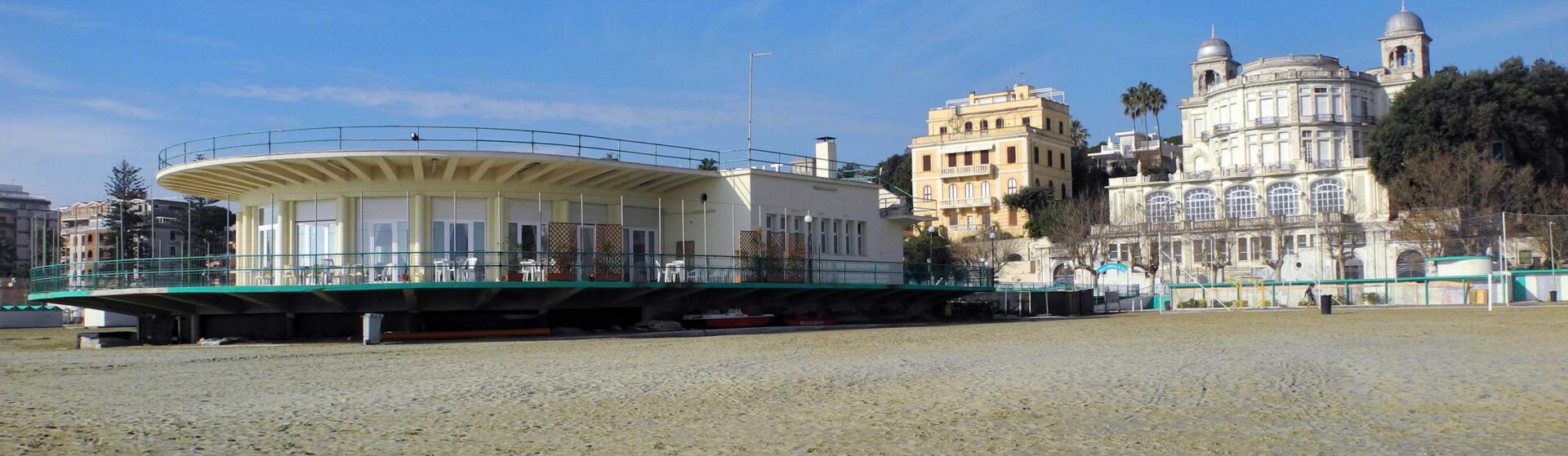 Modern circular seaside café on stilts with glass walls, on a sandy beach, with historic domed buildings in the background.