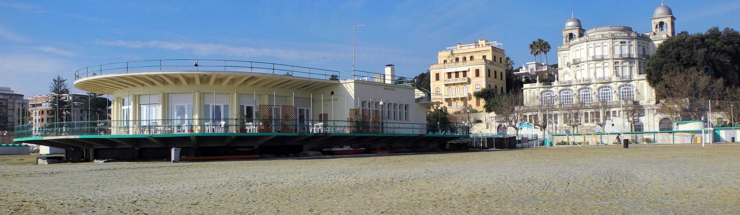 Modern circular seaside café on stilts with glass walls, on a sandy beach, with historic domed buildings in the background.