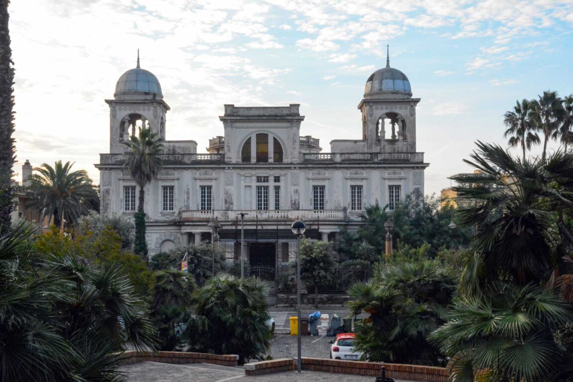 Historic white villa with two domed towers, arched windows, surrounded by lush palm trees.