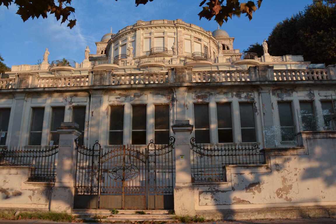 Dilapidated grand mansion with peeling white façade, tall windows, statues along the balcony, and a rusted iron gate.