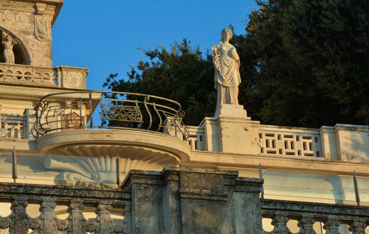 Classical statue on a stone pedestal atop a terrace with ornate wrought-iron railing, blue sky and trees.