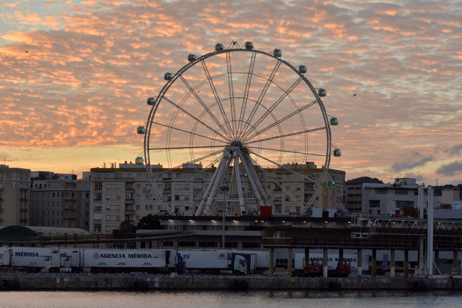 Malaga. Ruota panoramica al tramonto sul porto con skyline di edifici e camion in banchina