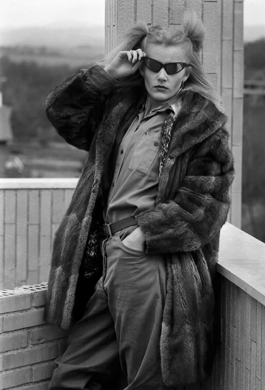 Black-and-white fashion photo: woman in a fur coat and sunglasses leaning on a rooftop railing.