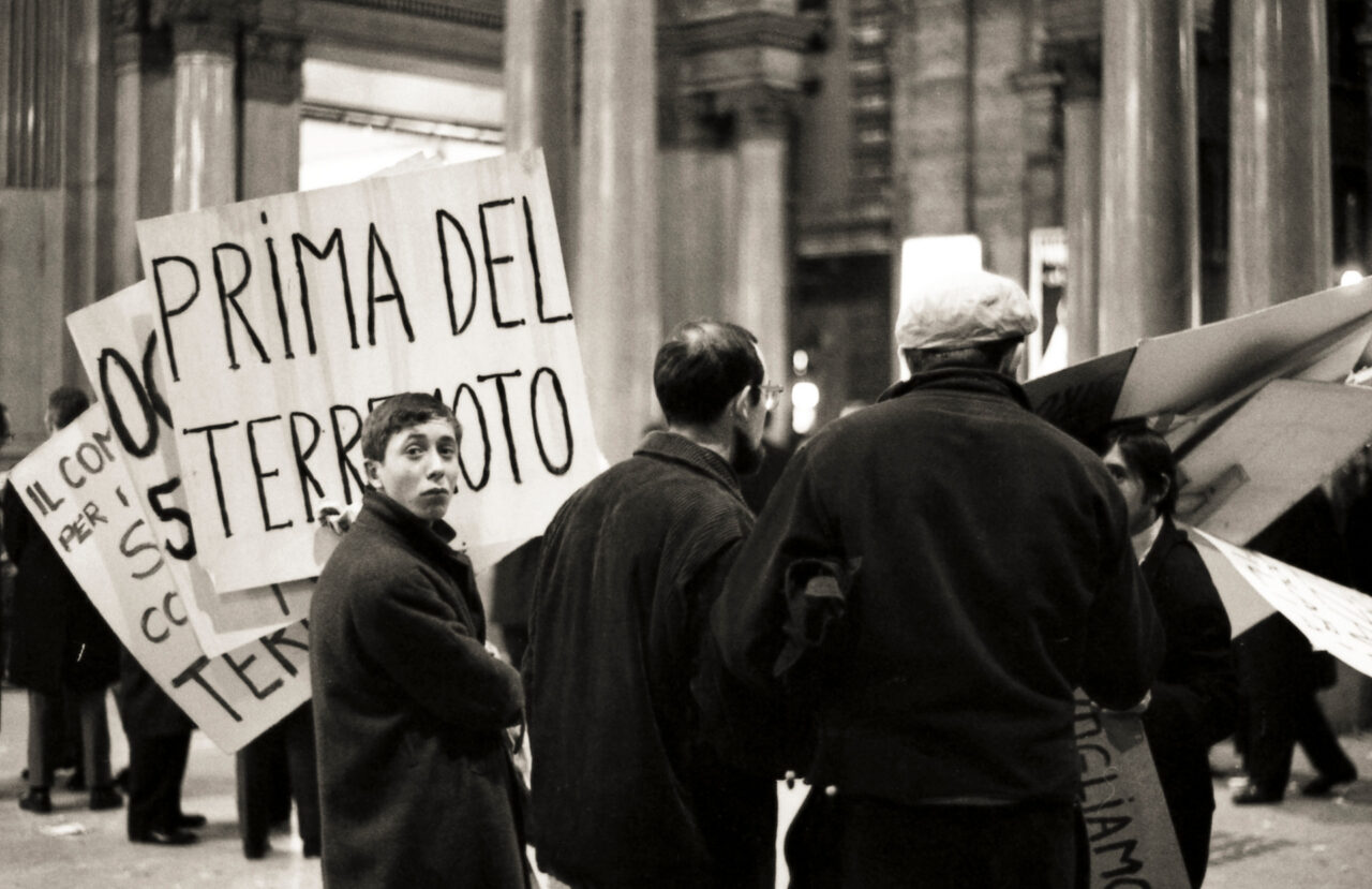 Black-and-white photo of protesters in a city, holding handwritten signs, including Prima del terremoto, at night.