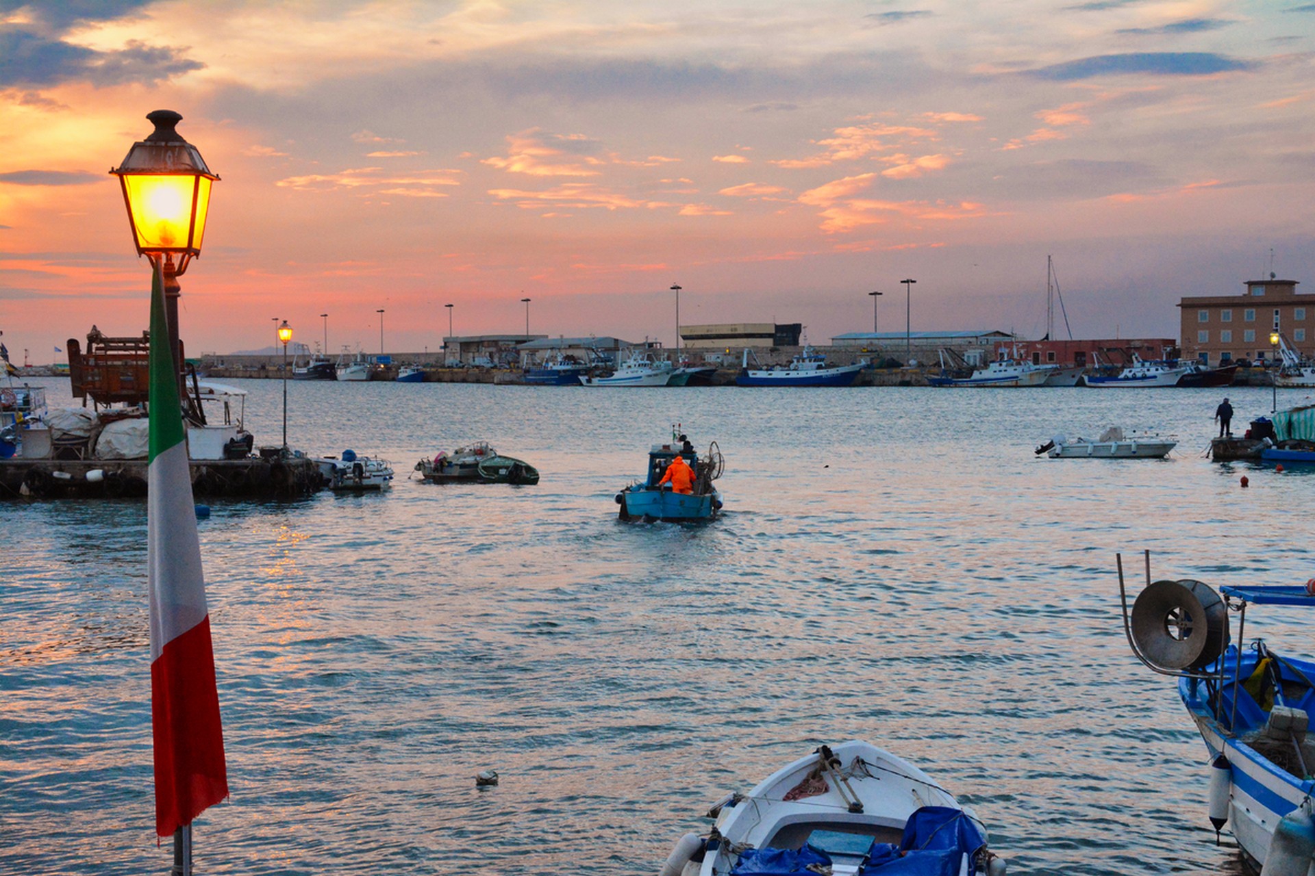 Sunset harbor scene with boats, calm water, a glowing lamp, and an Italian flag in the foreground.
