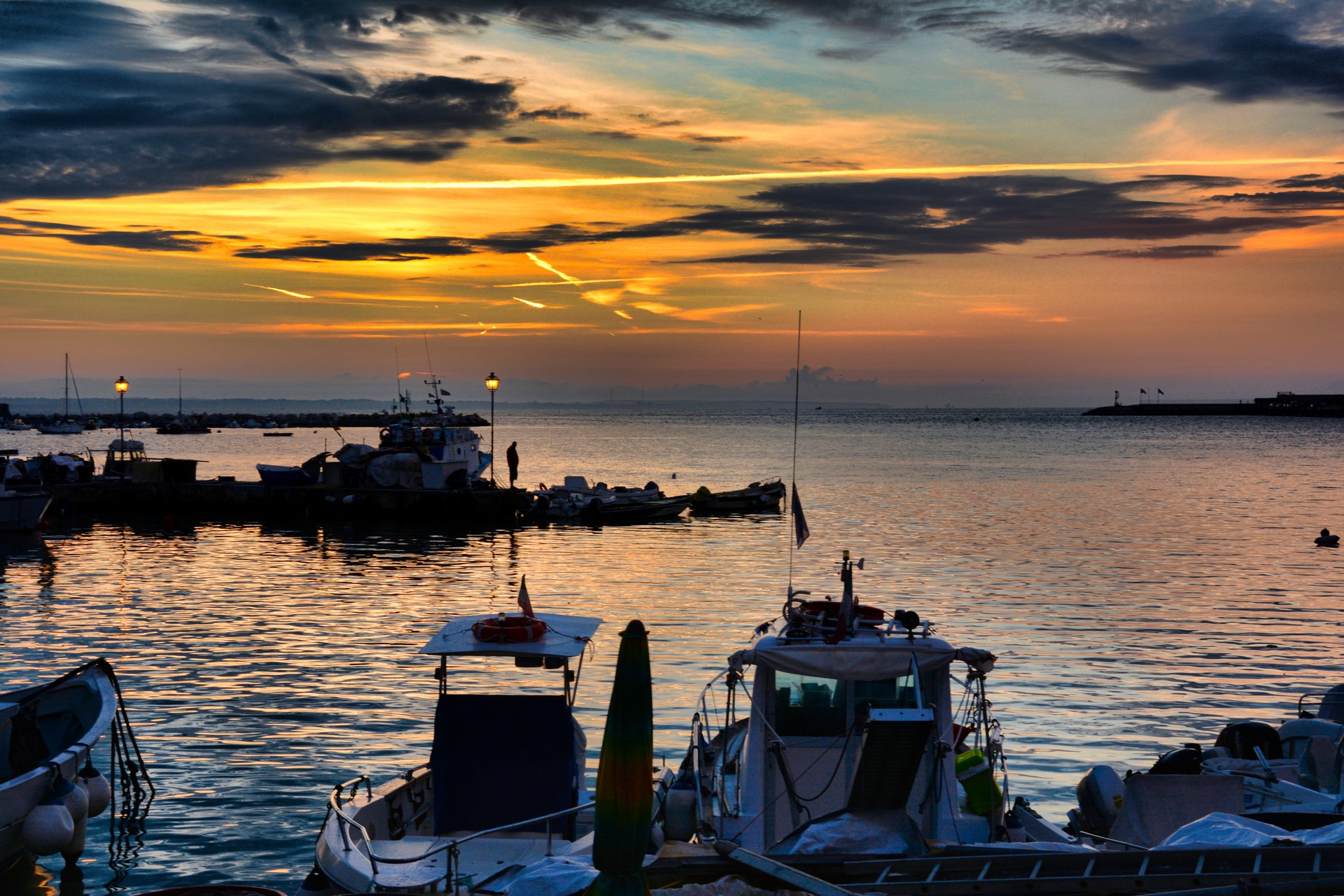 Sunset over a tranquil harbor with moored boats and silhouetted figures reflected in orange water.