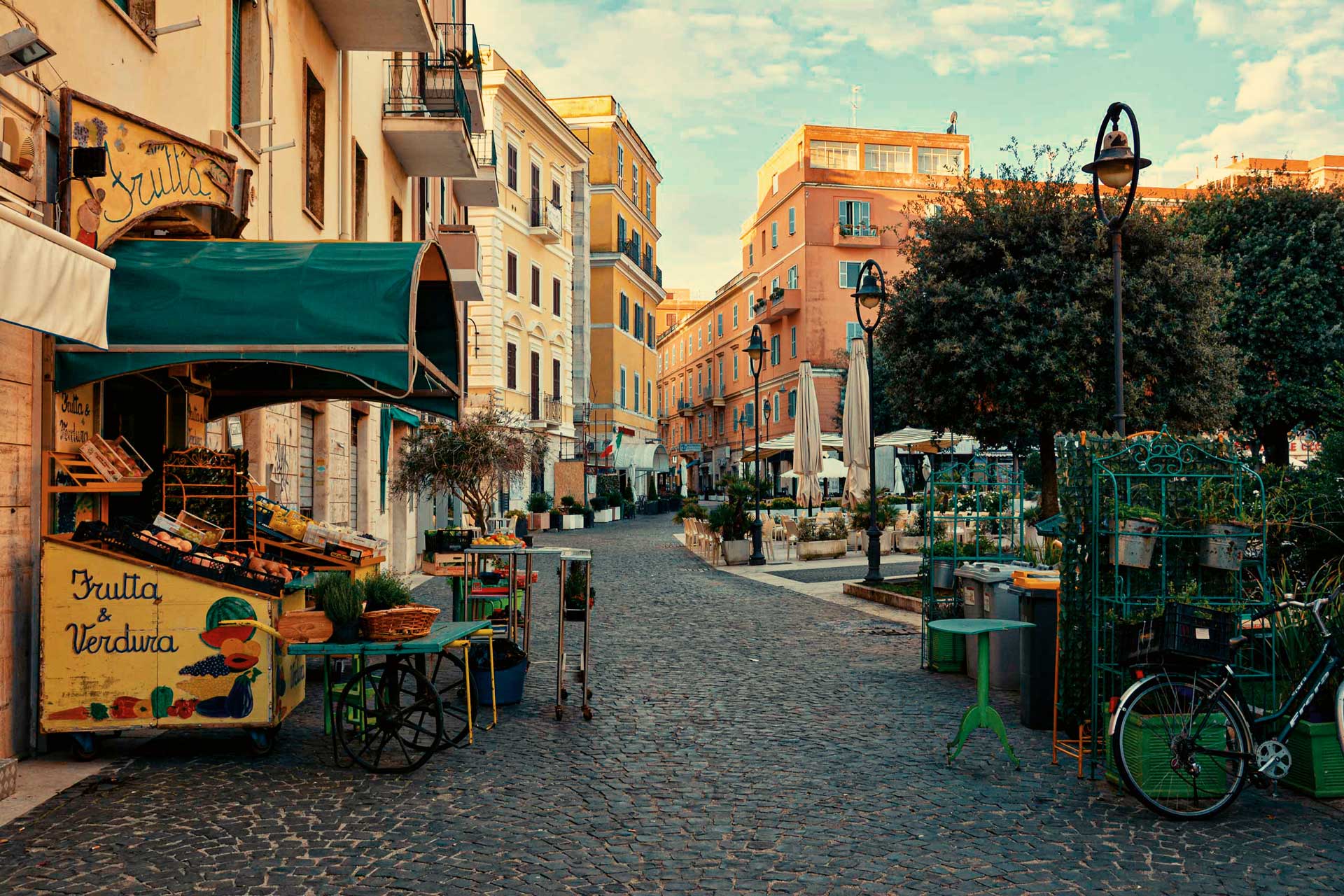 Sunny Italian street scene with cobblestone road, fruit stall, pastel buildings, outdoor seating, and bicycles.