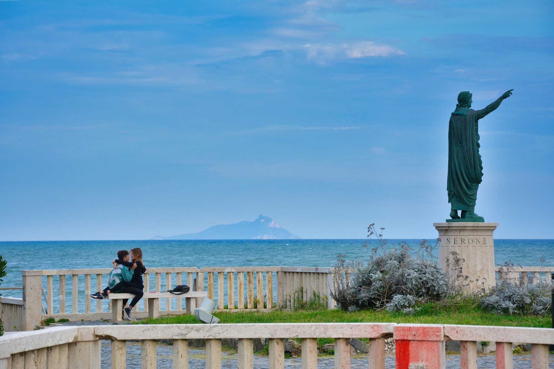 Couple hugging on a seaside bench near a Nerone statue, pointing toward the sea, with an island on the distant horizon.