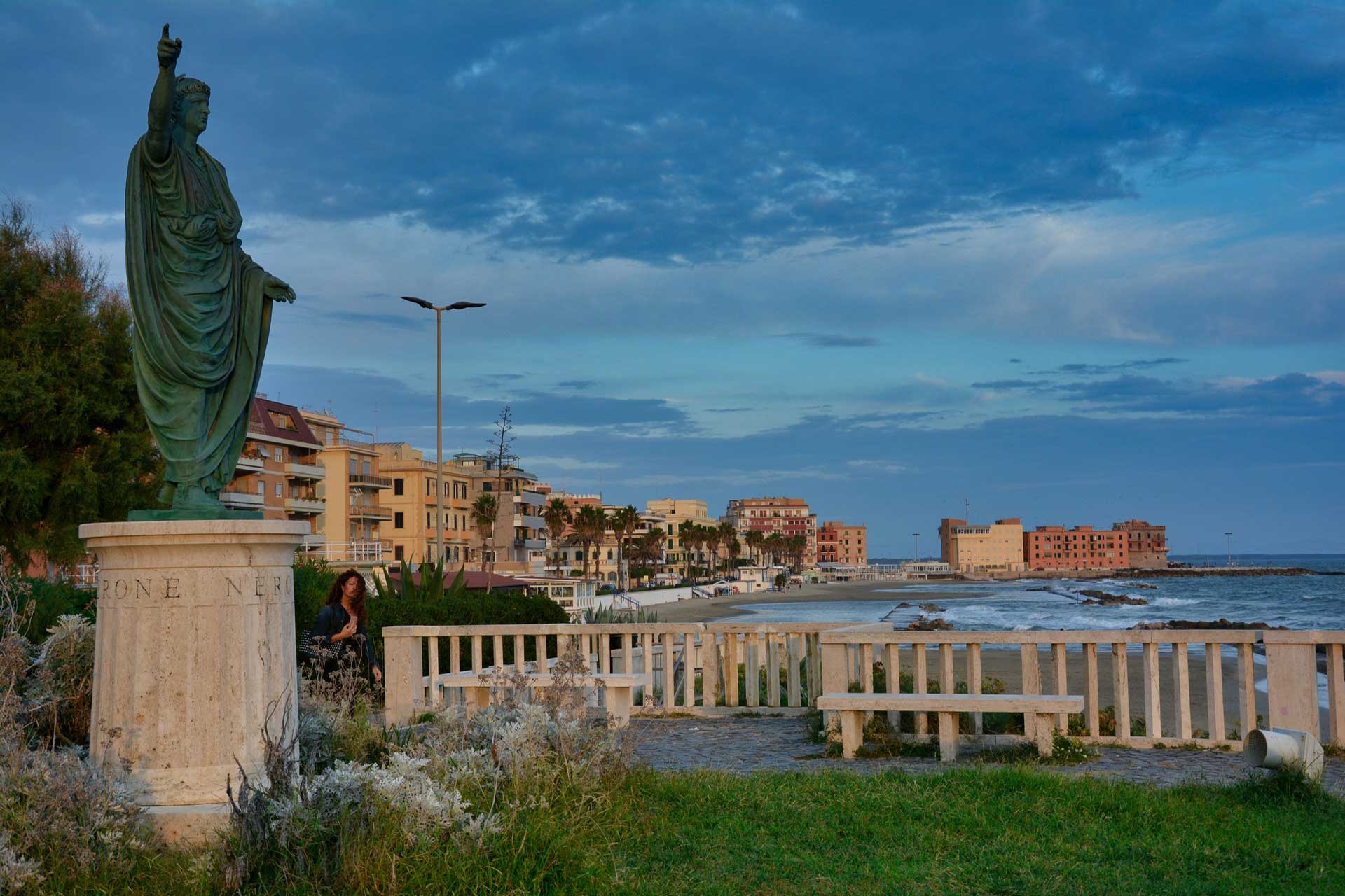 Bronze statue with raised arm on a pedestal beside a seaside promenade, overlooking pastel buildings and the ocean.