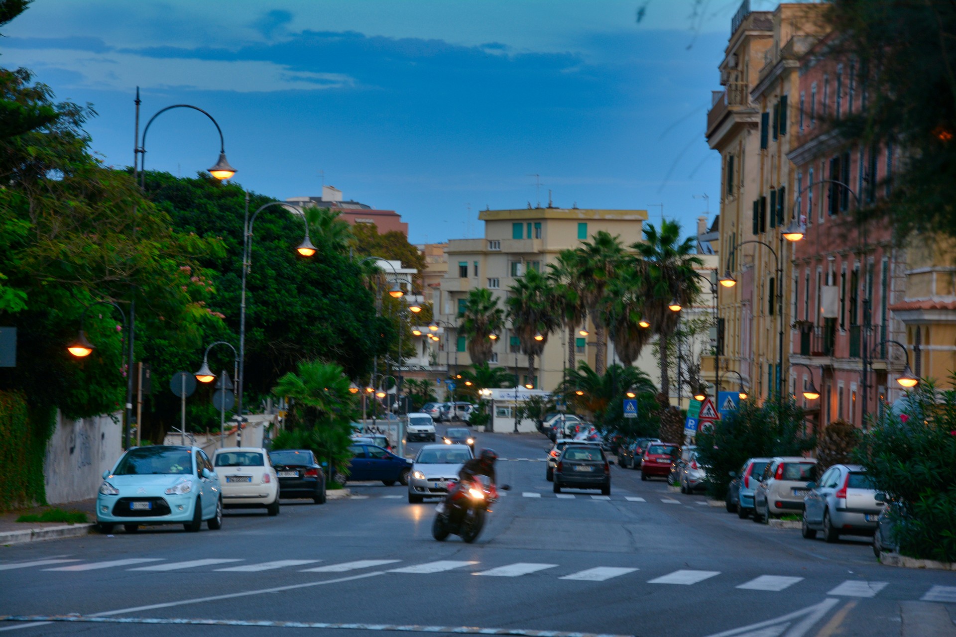 Twilight street scene in a Mediterranean town: palm-lined avenue, pastel buildings, parked cars, and a moving motorcycle.