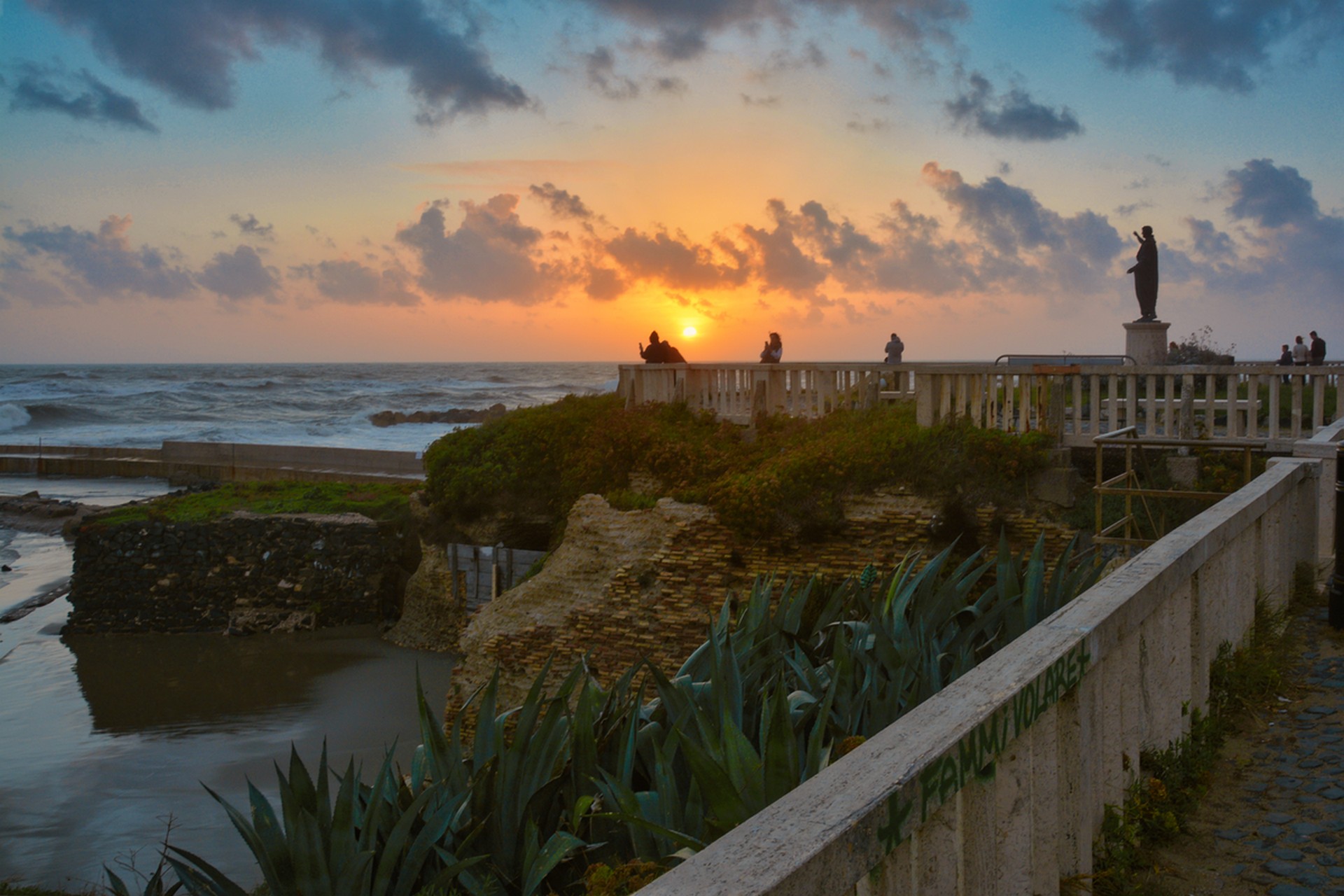 Sunset over a coastal promenade with a statue, stone railings, and visitors near the sea.