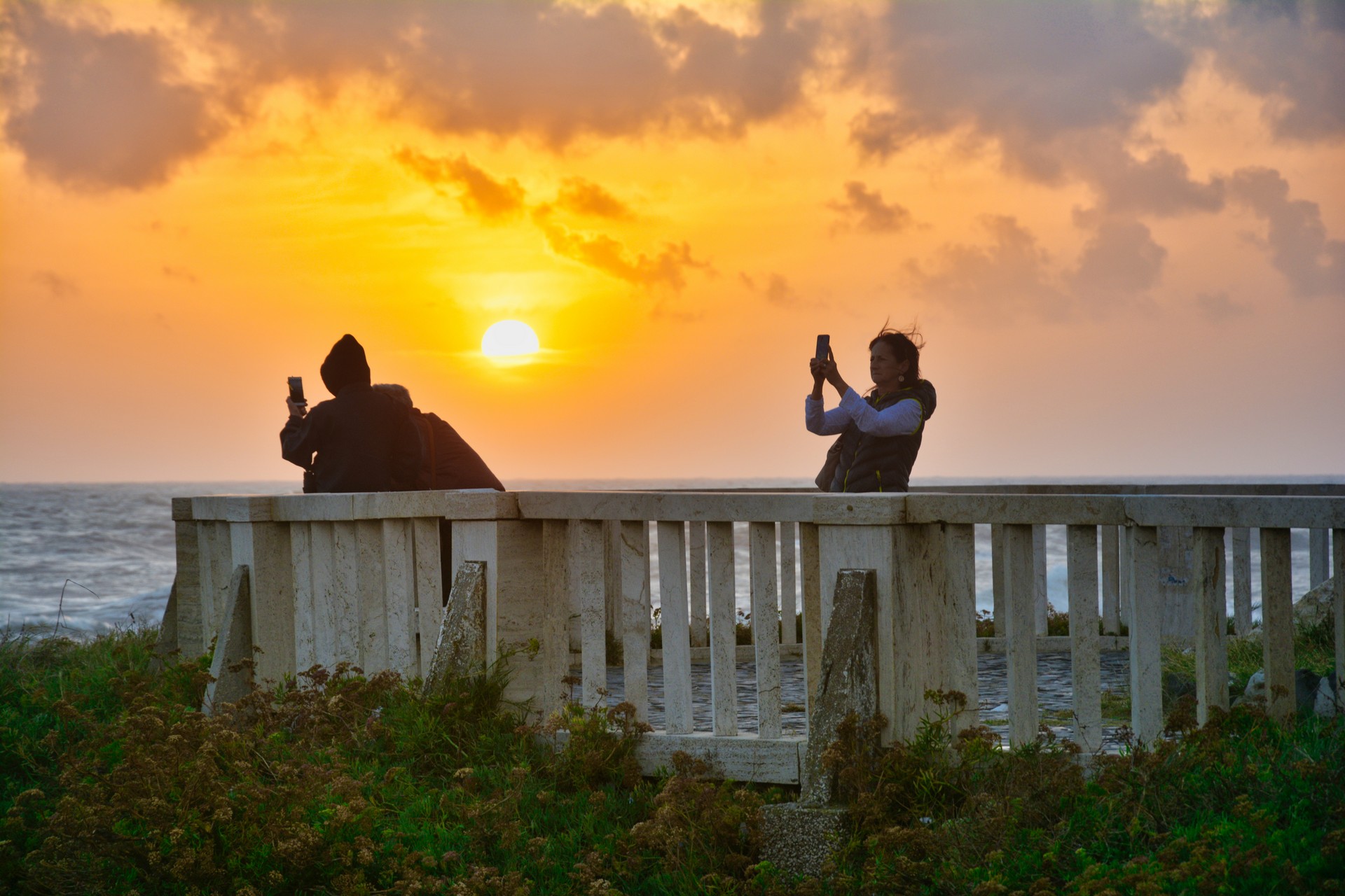 Two people on a seaside white concrete railing at sunset, taking photos with their phones as the sun dips over the ocean.