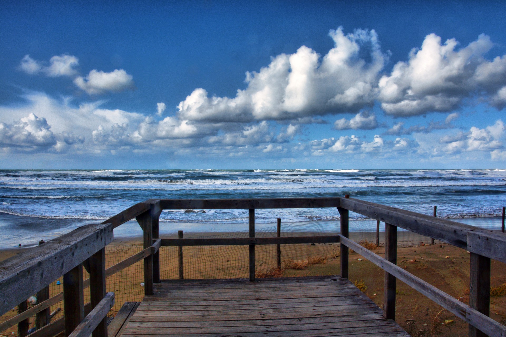 Weathered wooden boardwalk leading to a blue ocean beach under a dramatic cloudy sky.