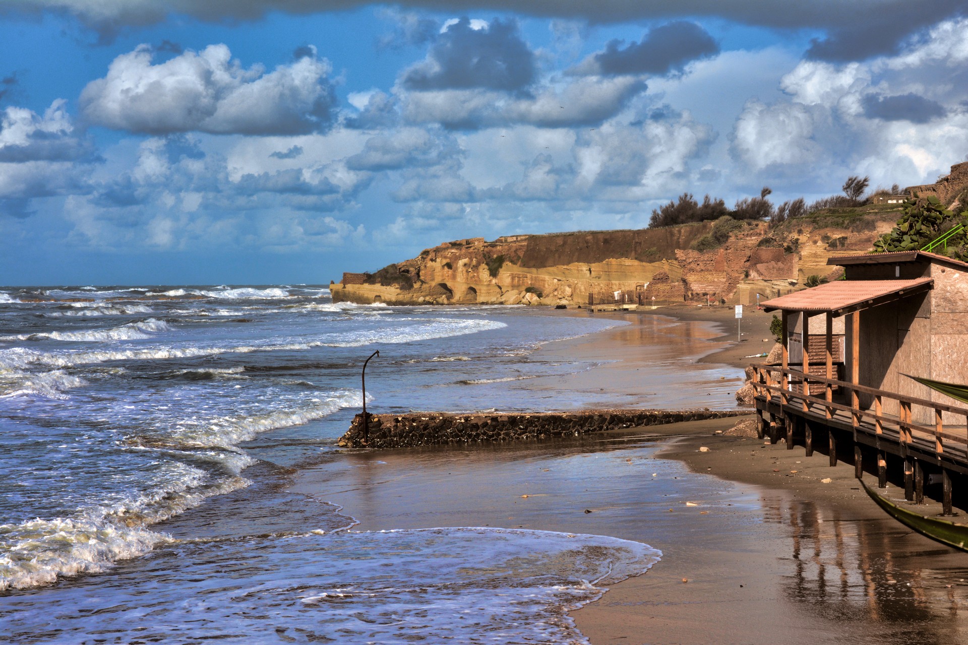 Sunny coastal scene with rocky cliffs, sandy beach, crashing waves, and a wooden boardwalk along the shore.