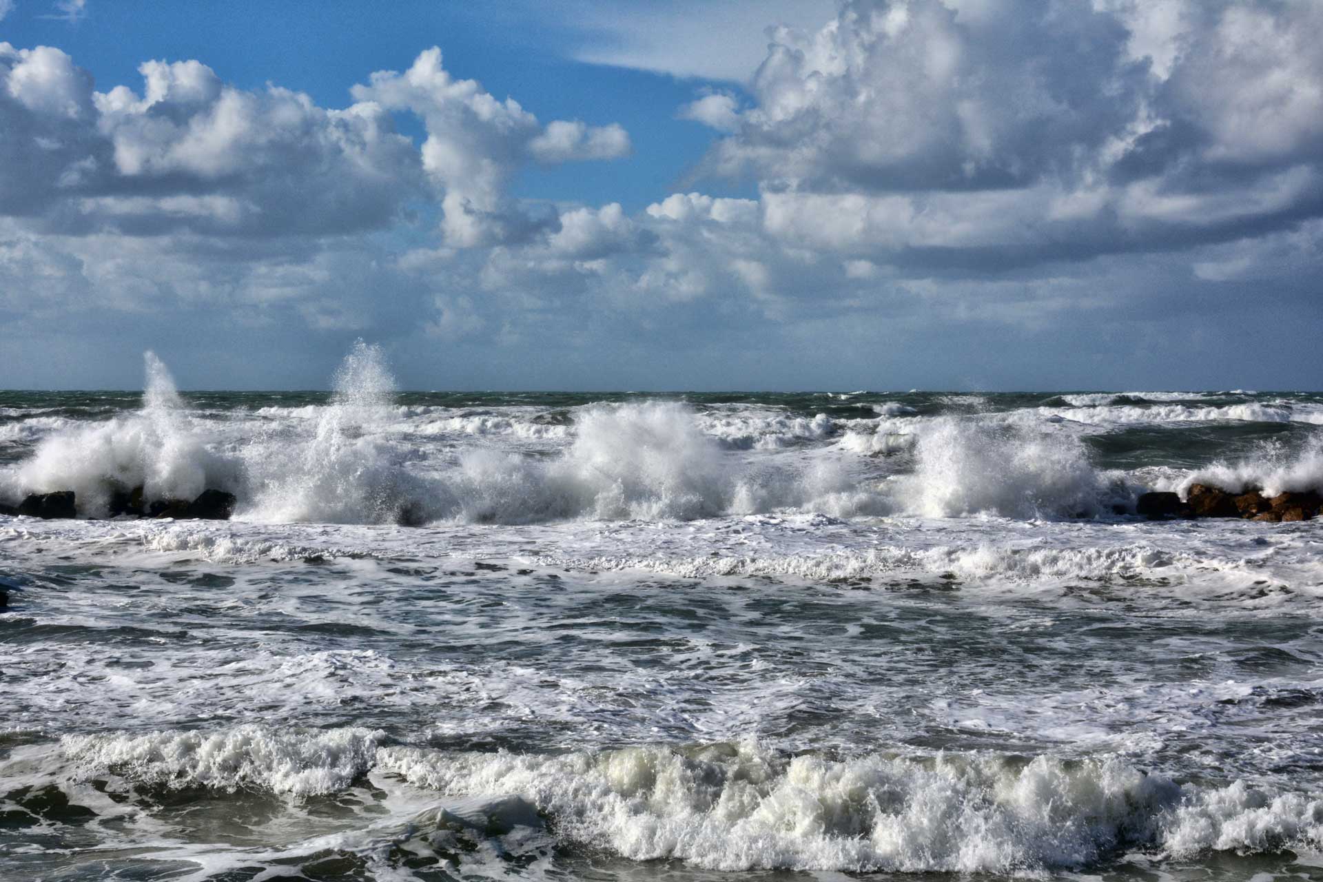 Coastal scene with rough ocean waves crashing on a rocky shoreline under a cloudy blue sky.