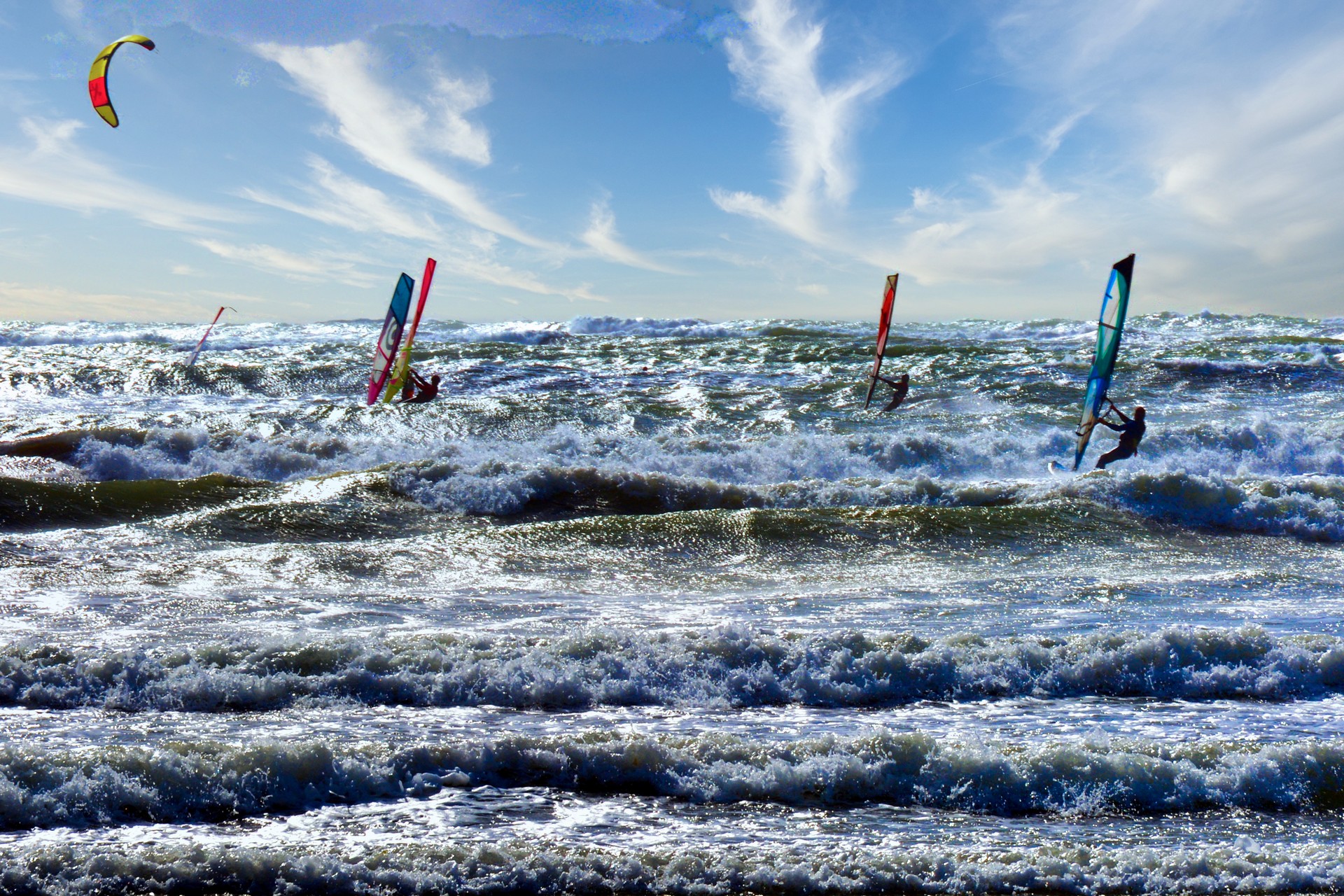 Wind surfers ride tall waves under a blue sky with colorful sails.