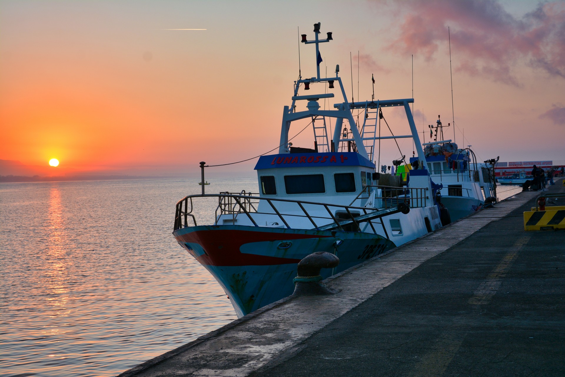 Sunset over a harbor with moored fishing boats along a concrete pier and calm water reflecting orange sky.