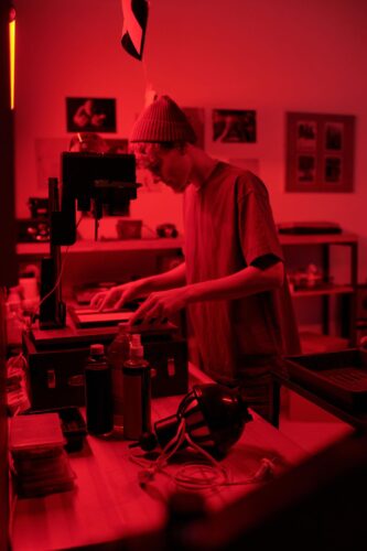 Young man in a beanie producing music in a red-lit home studio with mixer, keyboard, and headphones.