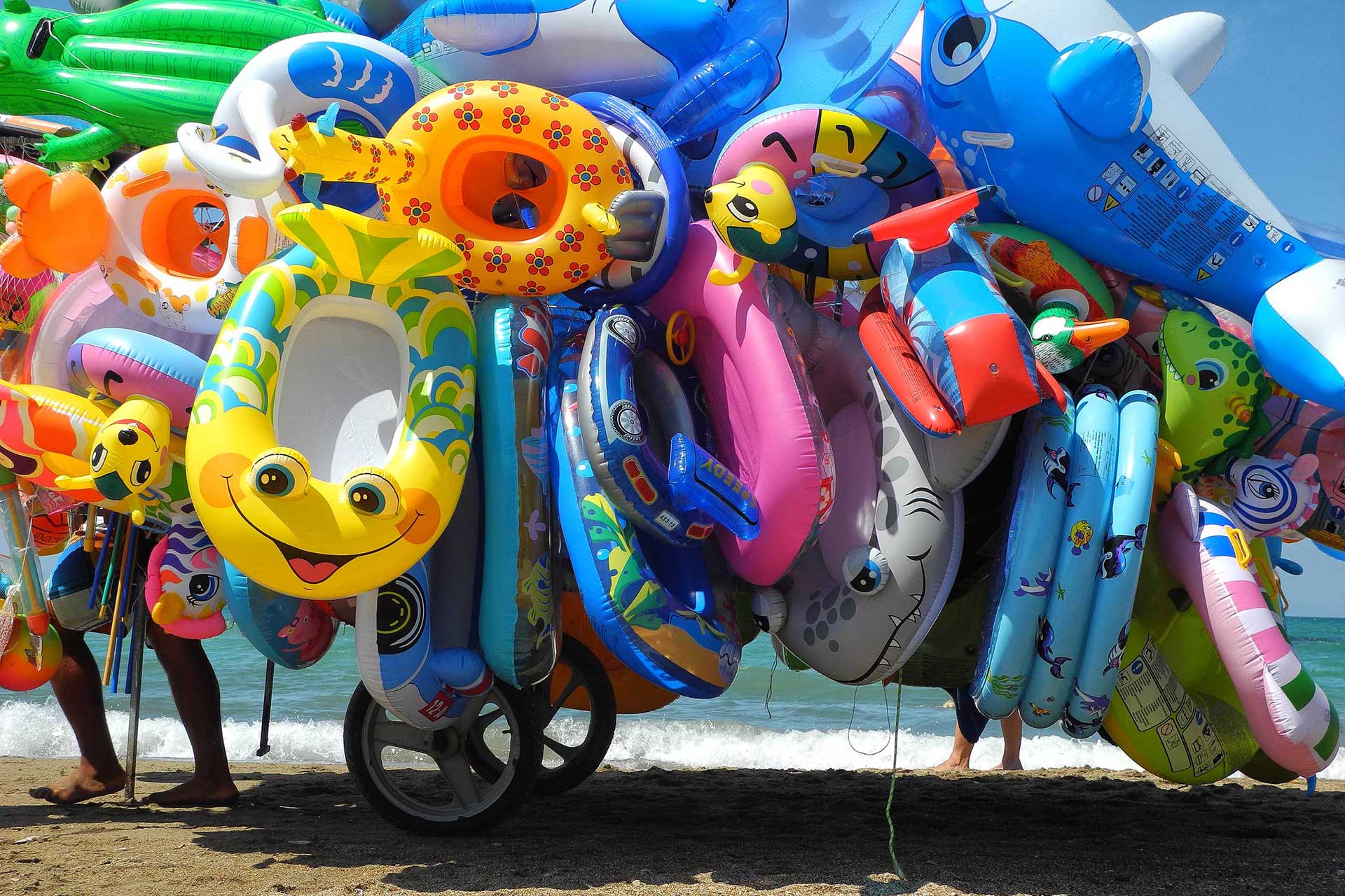 Colorful inflatable pool floats with cartoon faces piled on a sunny beach by the sea.